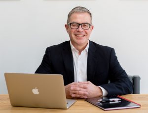 Photograph of Non-Executive Director, Tim Davies. He is sitting at a desk, smiling and facing the camera in a business suit. There is an Apple MacBook and notebook on the desk.