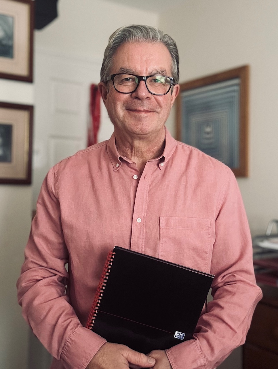 Tim Davies standing in his home office, wearing a pink shirt, carrying a notebook, smiling into the camera